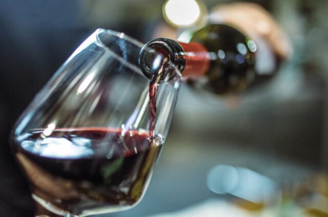 Unrecognizable person, holding a bottle of red wine, is pouring some red wine in a wine glass.